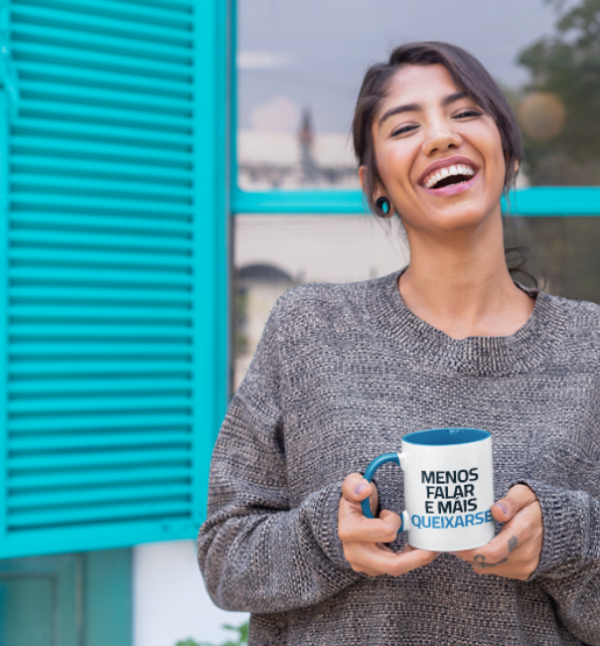 Una mujer sonriente sostiene una taza con texto cerca de una ventana con persianas azules.
