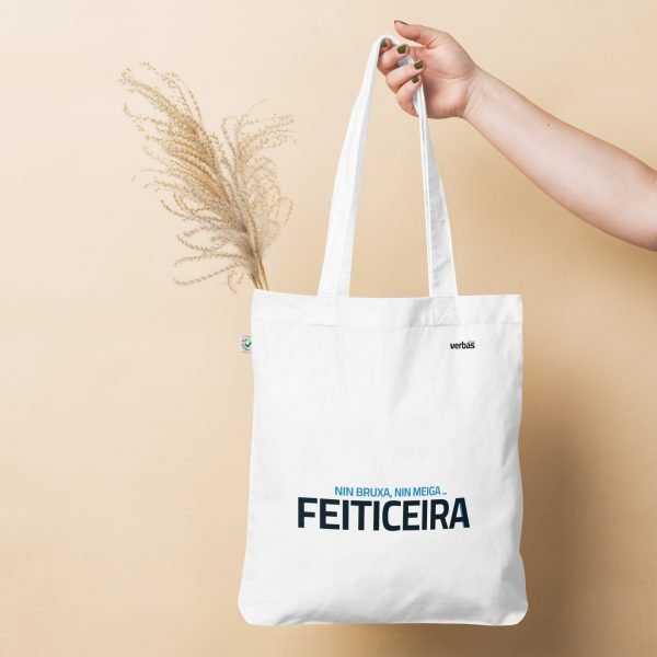 Una mano sostiene una bolsa de tela con la palabra FEITICEIRA impresa, sobre un fondo beige.